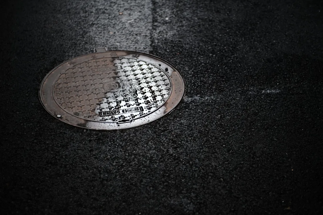 Close-up of a wet manhole cover on a rainy urban street, reflecting light. Photo by YWEN ZHU on Pexels