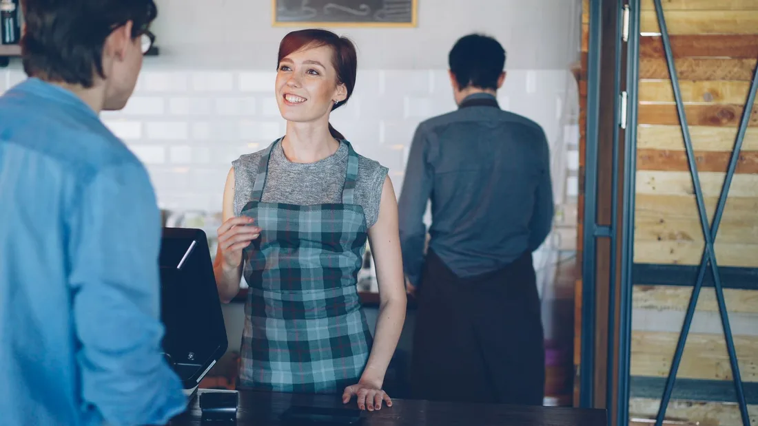 Smiling barista at the counter engaging with a customer in a cozy café. Photo by Vitaly Gariev on Pexels