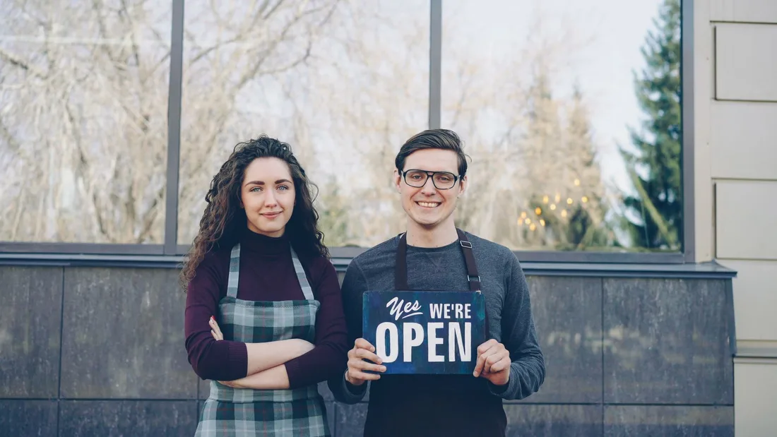 Two business owners standing outside, holding a 'Yes We're Open' sign, welcoming customers. Photo by Vitaly Gariev on Pexels