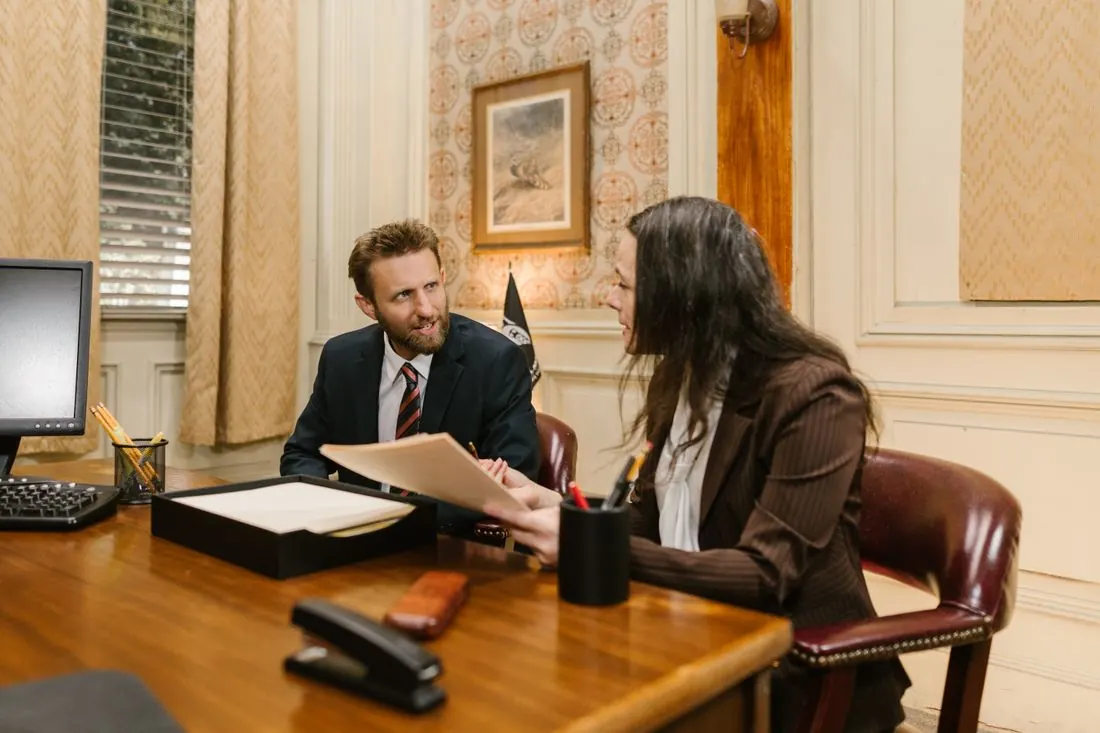 Man in a suit and tie and a woman in a brown blazer reading a printed document across a wooden desk with a desktop monitor and a stapler. Photo by RDNE Stock project on Pexels