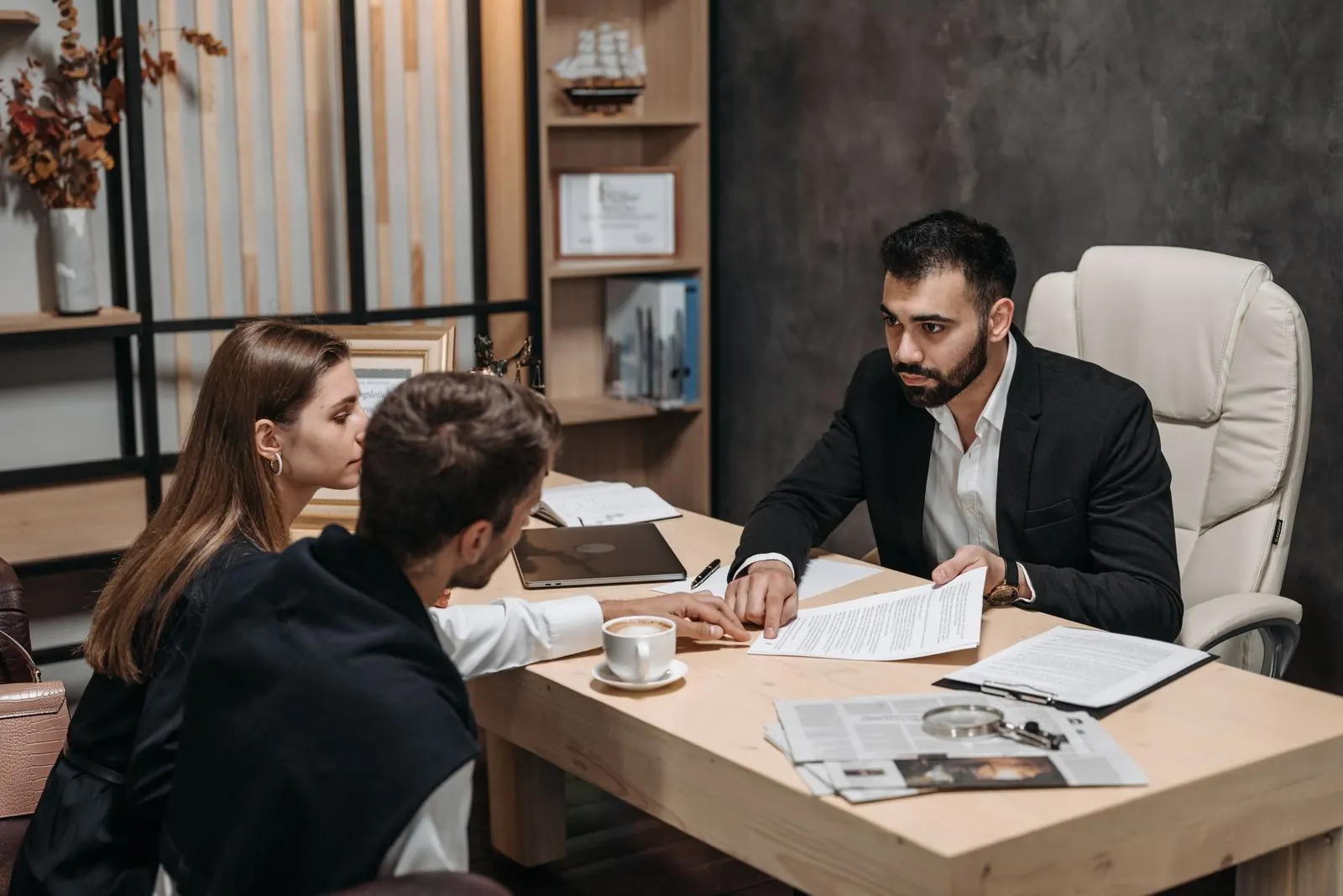 Man in a dark suit at a desk holding a printed document while a man and a woman seated across from him look at the page, with a coffee cup, a clipboard and a newspaper on the desk. Photo by Pavel Danilyuk on Pexels