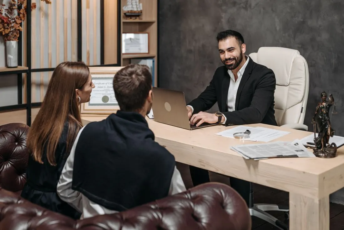 Smiling man in a dark suit typing on a laptop at a desk while a man and a woman sit across from him on a leather sofa, with a framed certificate and a Lady Justice statuette nearby. Photo by Pavel Danilyuk on Pexels