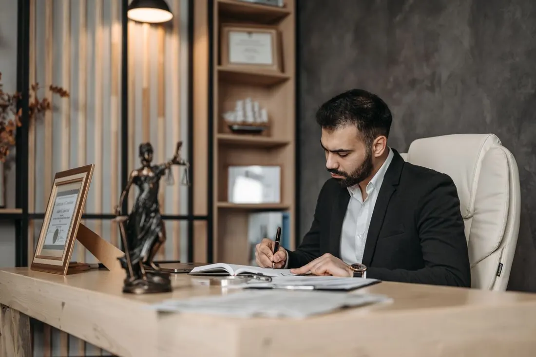 Bearded man in a dark suit writing in an open notebook at a wooden desk, with a Lady Justice statuette and a framed certificate beside him. Photo by Pavel Danilyuk on Pexels