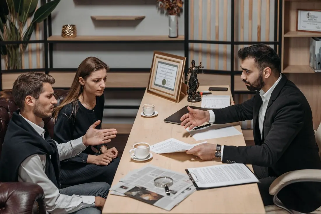 Man in a dark suit handing a printed page across a desk to a man and a woman seated on a leather sofa, with coffee cups and a clipboard on the desk. Photo by Pavel Danilyuk on Pexels