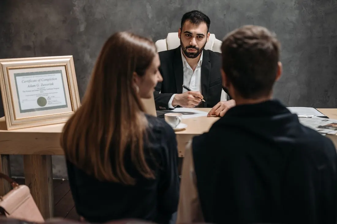 Man in a dark suit holding a pen at a desk, facing a woman and a man seen from behind, with a framed certificate and a coffee cup on the desk. Photo by Pavel Danilyuk on Pexels