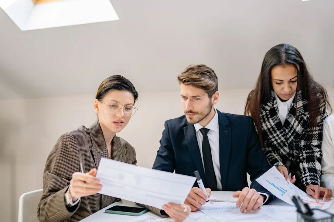 Three colleagues at a white table reviewing printed pages, one holding a sheet with charts and another holding a page with a line graph. Photo by Yan Krukau on Pexels