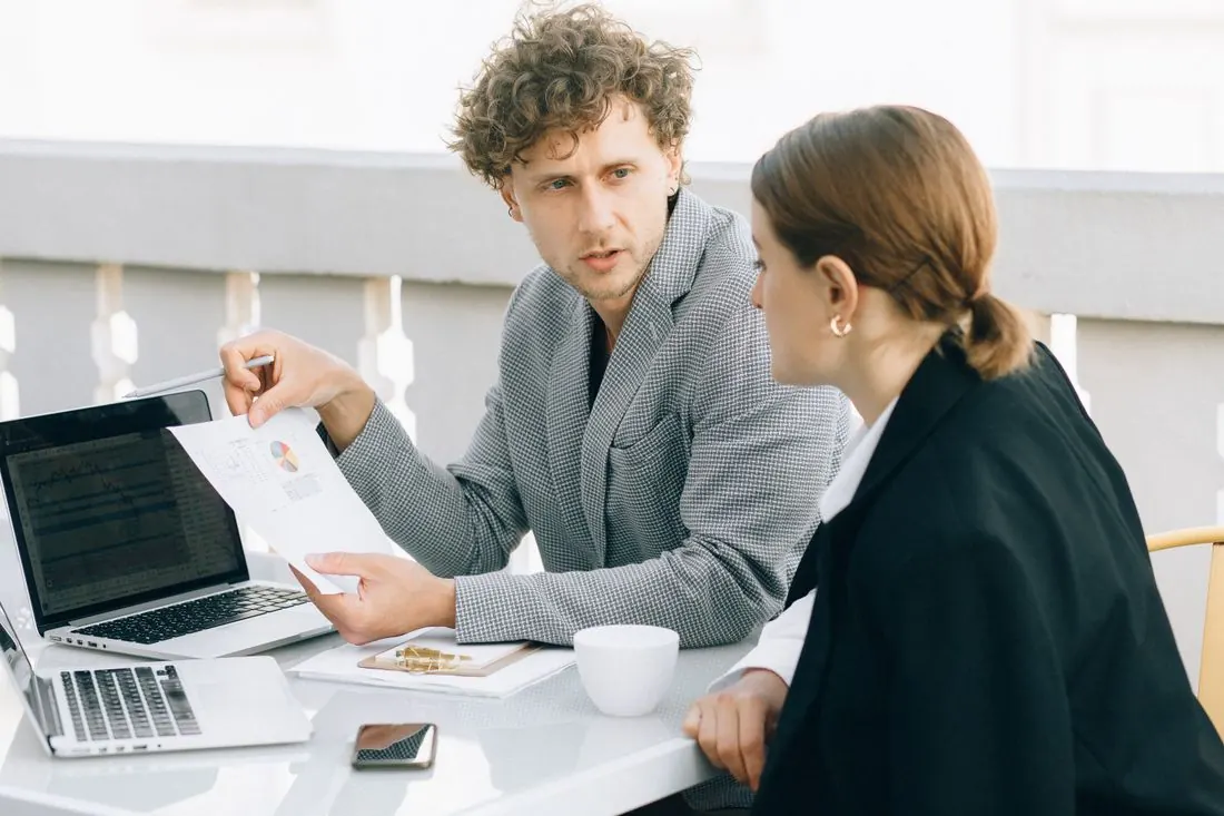 Man in a grey checked jacket holding a printed chart beside a woman in a black blazer, with an open laptop and a coffee cup on a white table outdoors. Photo by Nataliya Vaitkevich on Pexels