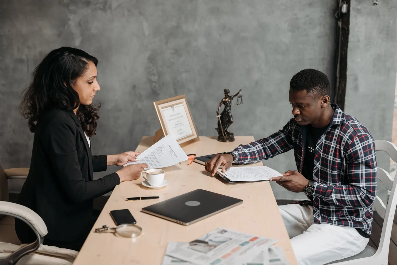 Woman in a black blazer and a man in a checked shirt reading printed documents across a desk with a laptop and a framed certificate. Photo by Pavel Danilyuk on Pexels