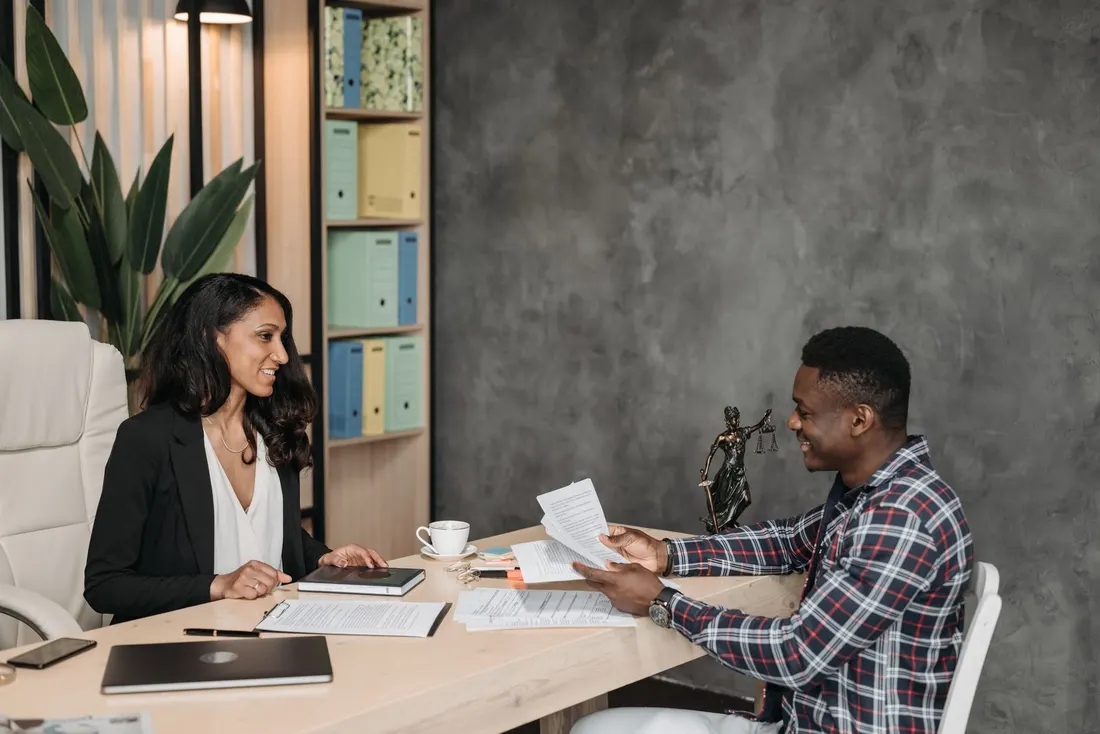 Woman in a black blazer smiling across a desk at a man in a checked shirt holding papers, with a laptop, coffee cup and box files behind. Photo by Pavel Danilyuk on Pexels