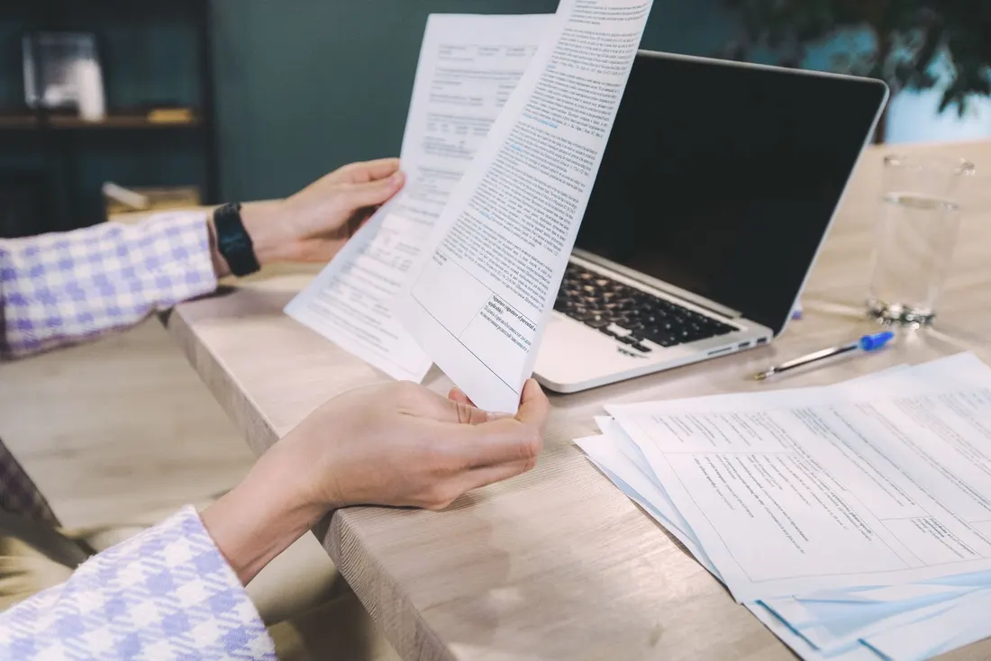 Hands holding printed pages next to an open laptop, a pen and a stack of documents on a wooden desk. Photo by SHVETS production on Pexels