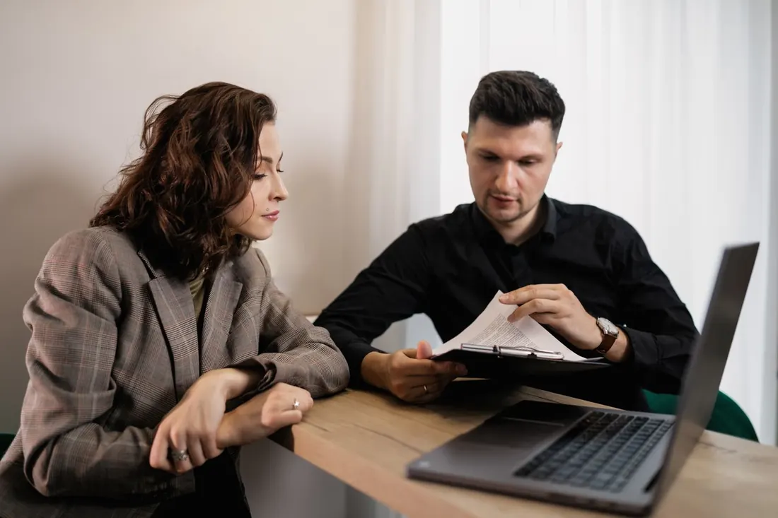 A woman in a plaid blazer and a man in a black shirt reviewing a printed page at a desk, with a laptop open. Photo by Alena Darmel on Pexels
