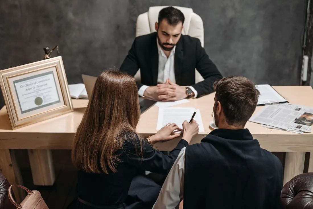 Man in a dark suit seated behind a desk while two clients sign a document, with a framed certificate on the desk. Photo by Pavel Danilyuk on Pexels