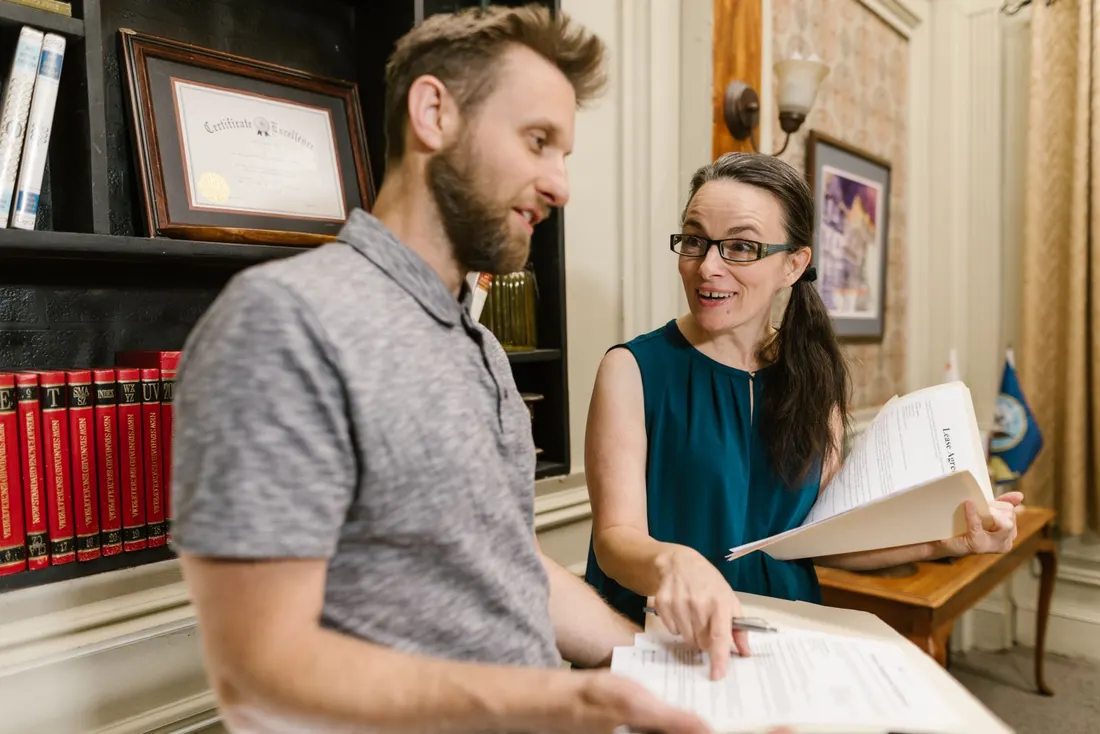 Woman in glasses holding a folder of papers talking with a man holding a document, in front of bookshelves. Photo by RDNE Stock project on Pexels