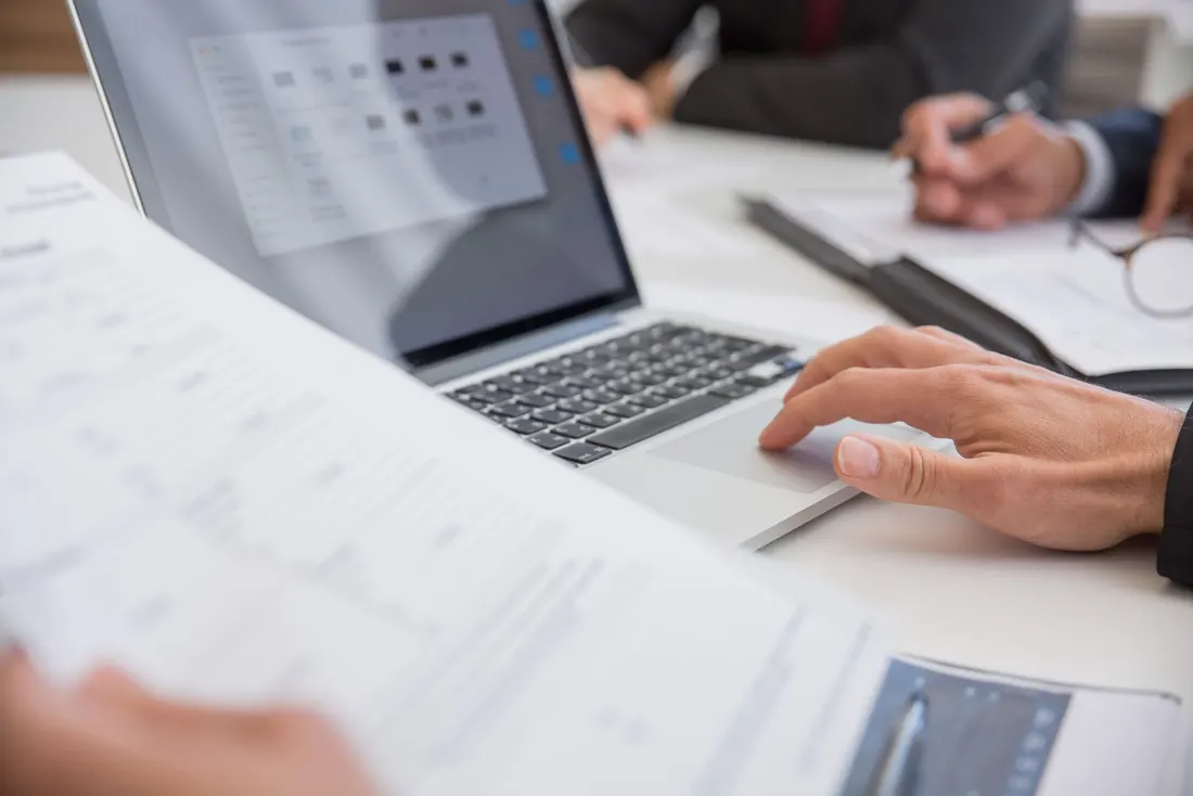 Close-up of a hand on a laptop trackpad with printed pages in the foreground and a colleague writing notes across the table. Photo by Kampus Production on Pexels