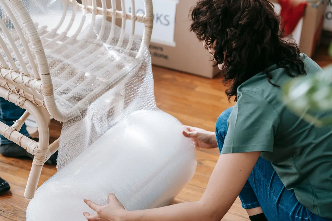 Woman kneeling to wrap a wicker chair with bubble wrap, preparing for relocation.. Photo by Blue Bird on Pexels