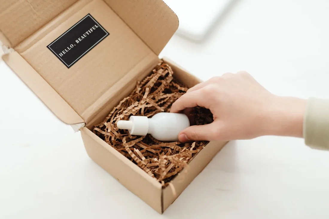 Hand unboxing a kraft box with a branded label and a bottle in paper filling. Photo by https://kaboompics.com/ on Pexels