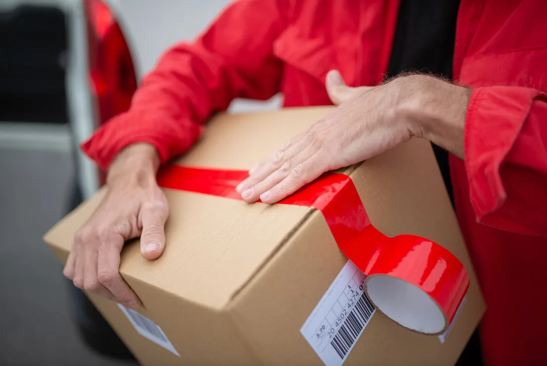 Person sealing a cardboard parcel with red packing tape and a barcode label. Photo by Kampus Production on Pexels