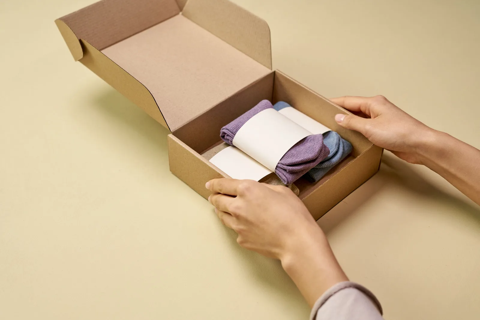 Hands placing folded socks into an open kraft mailer box on a cream background. Photo by Ron Lach on Pexels