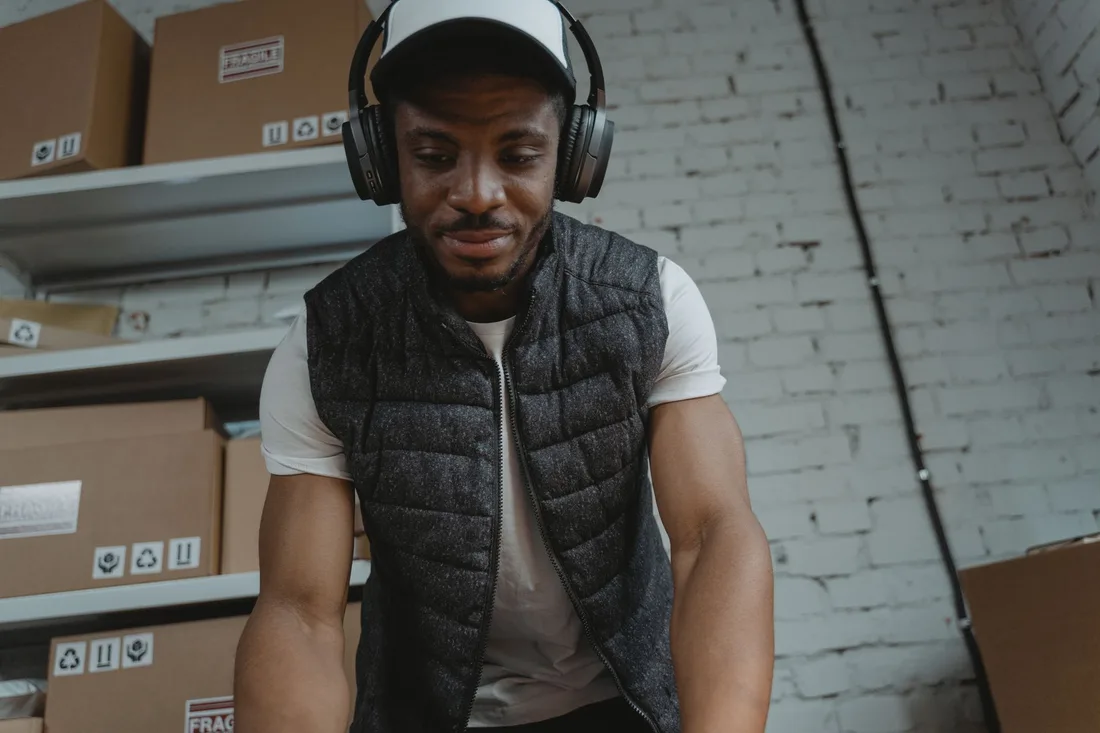 Smiling worker with headphones in a packing area with cardboard boxes on shelves. Photo by Tima Miroshnichenko on Pexels