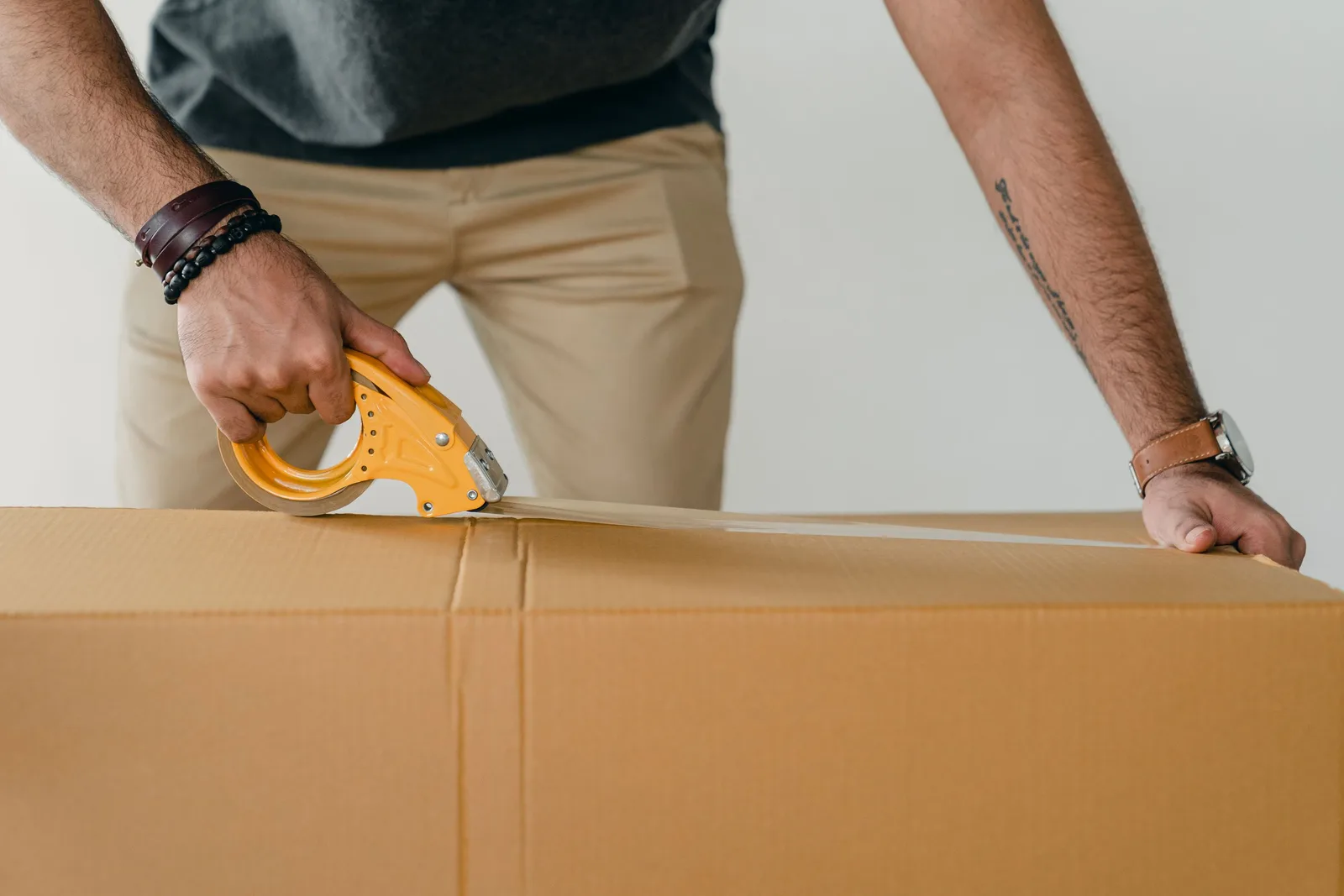 Crop anonymous young male packer in casual clothes using tape gun dispenser for sealing cardboard boxes. Photo by Ketut Subiyanto on Pexels