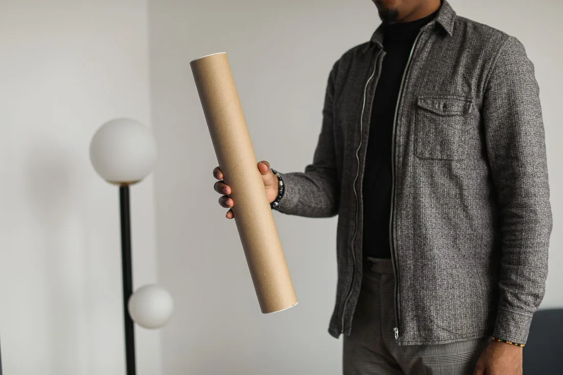 Man in a grey shirt holding a cardboard mailing tube indoors. Photo by Cup of  Couple on Pexels