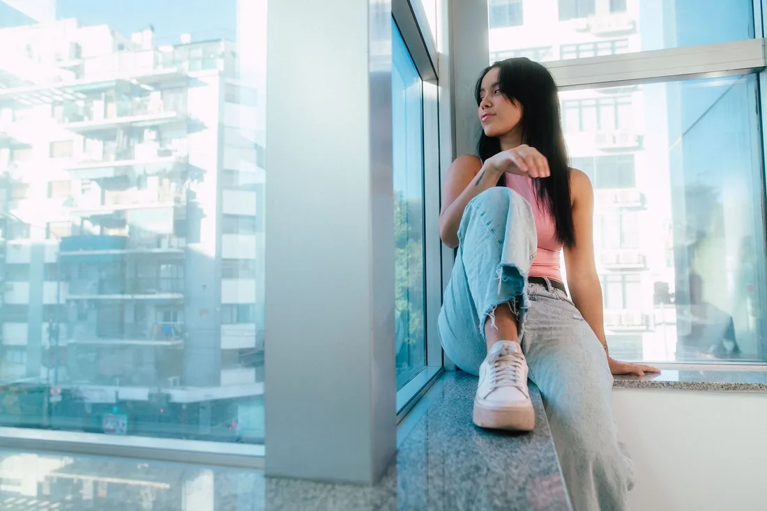 Young woman leaning on a large window in a city apartment with an urban view. Photo by Gera Cejas on Pexels