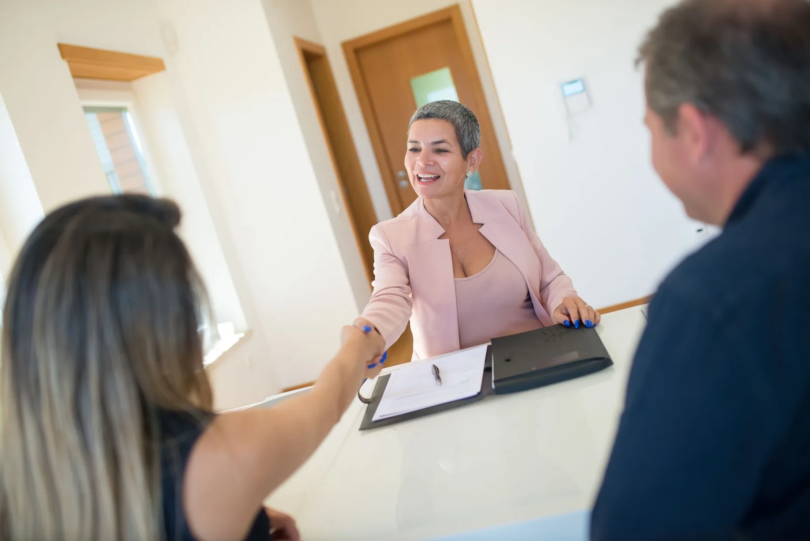 Real estate agent in pink blazer shaking hands with clients across a desk. Photo by Kampus Production on Pexels