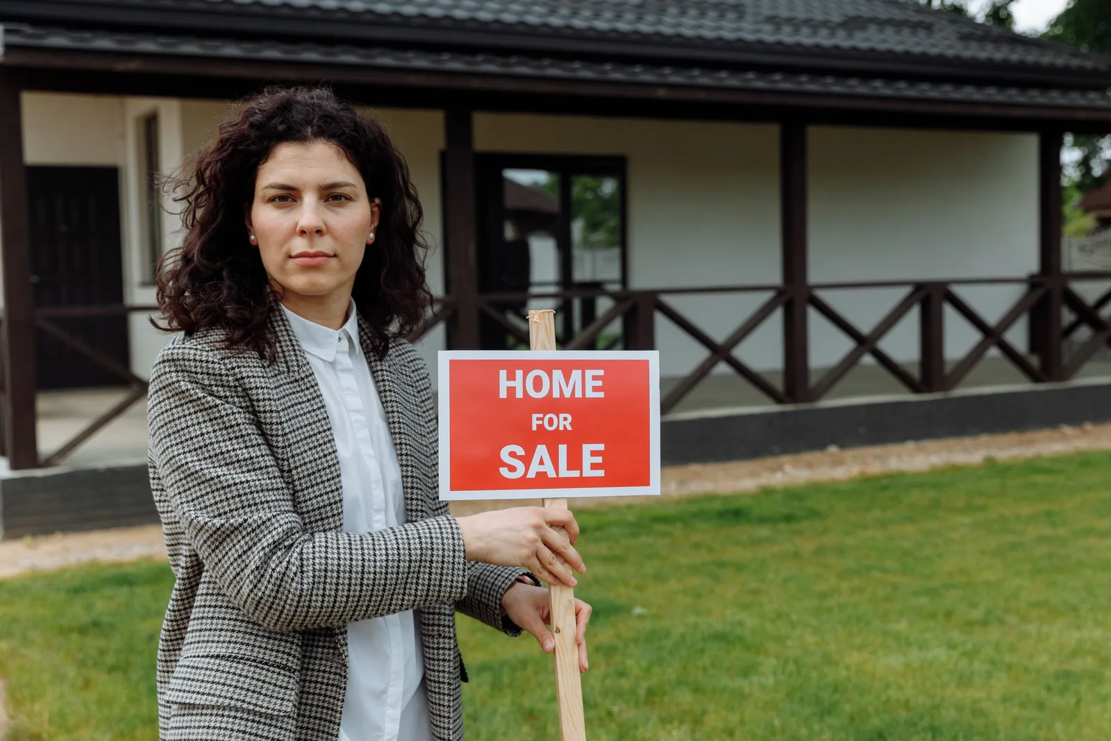 Real estate agent holding a home for sale sign in front of a single-storey house. Photo by Thirdman on Pexels