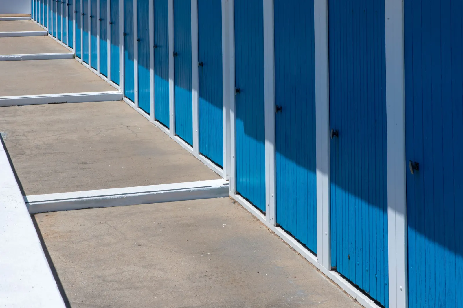A long row of bright blue roll-up storage unit doors with white frames along a concrete drive in strong sunlight. Photo by Jan van der Wolf on Pexels