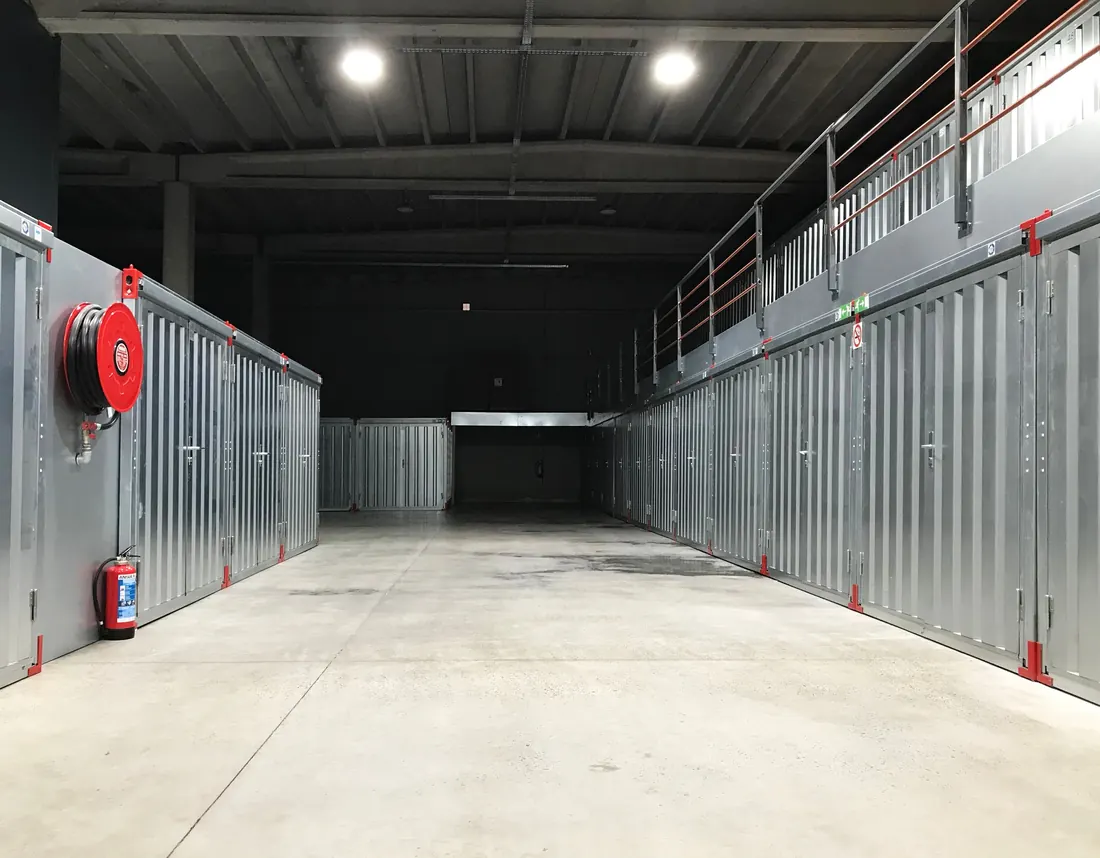 An indoor aisle of grey self-storage units with a mezzanine and railing on the right and a red fire-hose reel on the left wall. Photo by David Slaager on Pexels