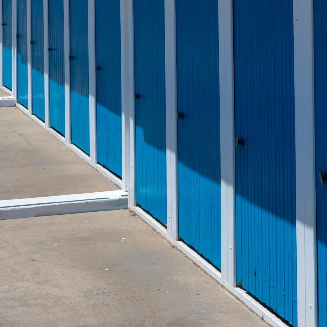 Close view along a row of bright blue roll-up storage unit doors with white frames beside a concrete drive. Photo by Jan van der Wolf on Pexels