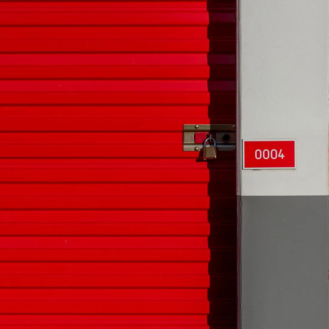 Close view of a red roll-up storage unit door with a padlock on its latch and a small plate reading 0004 on the pier beside it. Photo by Jan van der Wolf on Pexels