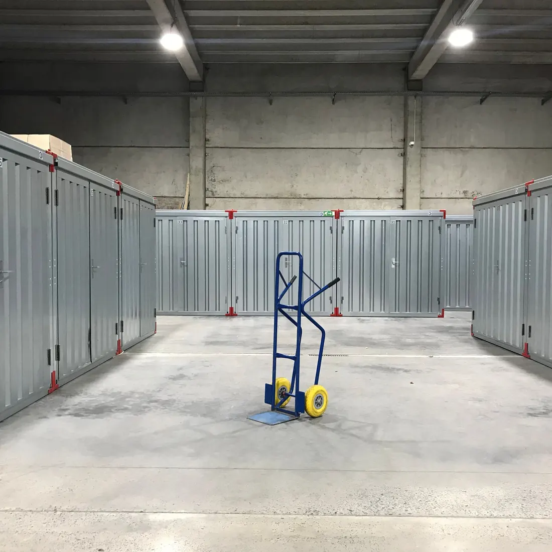 Indoor grey self-storage units around an open concrete floor, with a blue hand truck with yellow wheels standing in the aisle. Photo by David Slaager on Pexels