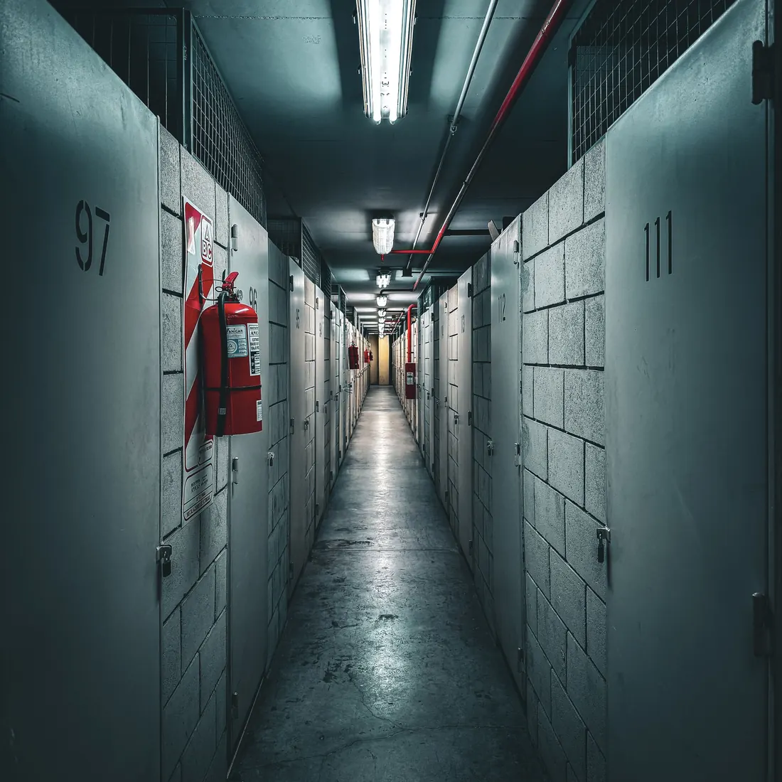 A narrow indoor storage corridor lined with numbered unit doors, including 97 and 111, with a red fire extinguisher on the left wall. Photo by Marcela Artola on Pexels
