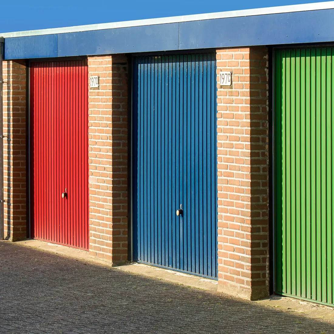 Red, blue and green roller doors on three storage units between brick piers, with small unit number plaques above the doors. Photo by Jan van der Wolf on Pexels