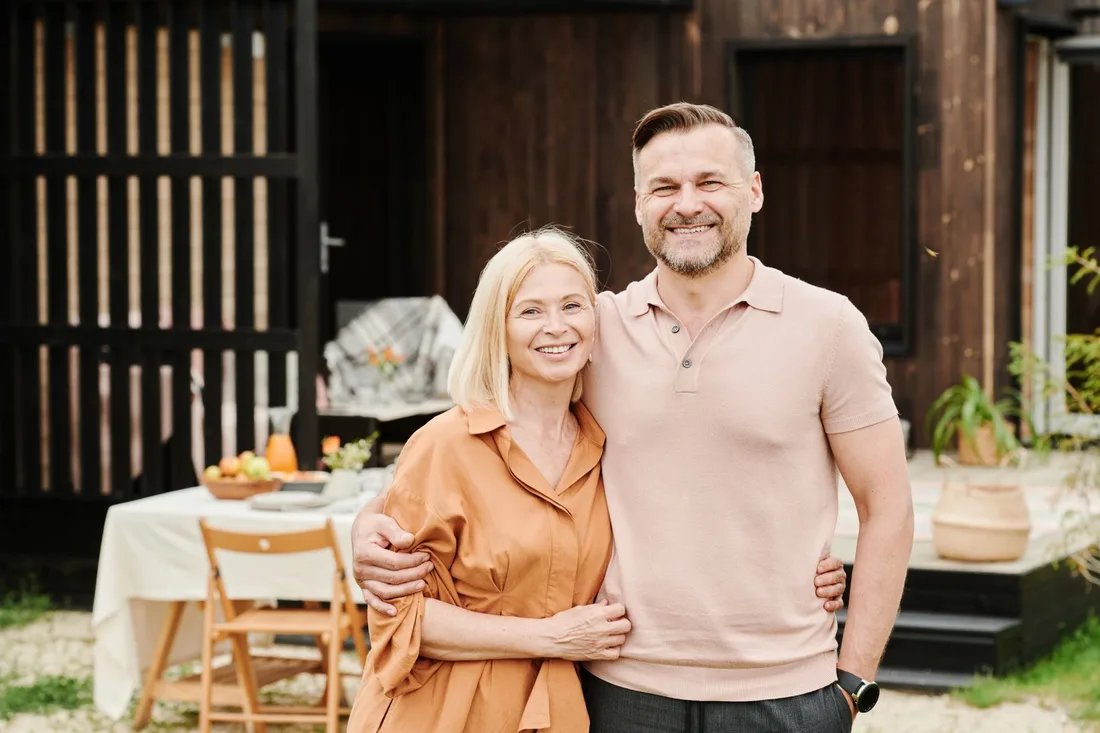 Smiling couple standing together outside a wooden holiday cabin with a table behind them. Photo by Julia M Cameron on Pexels