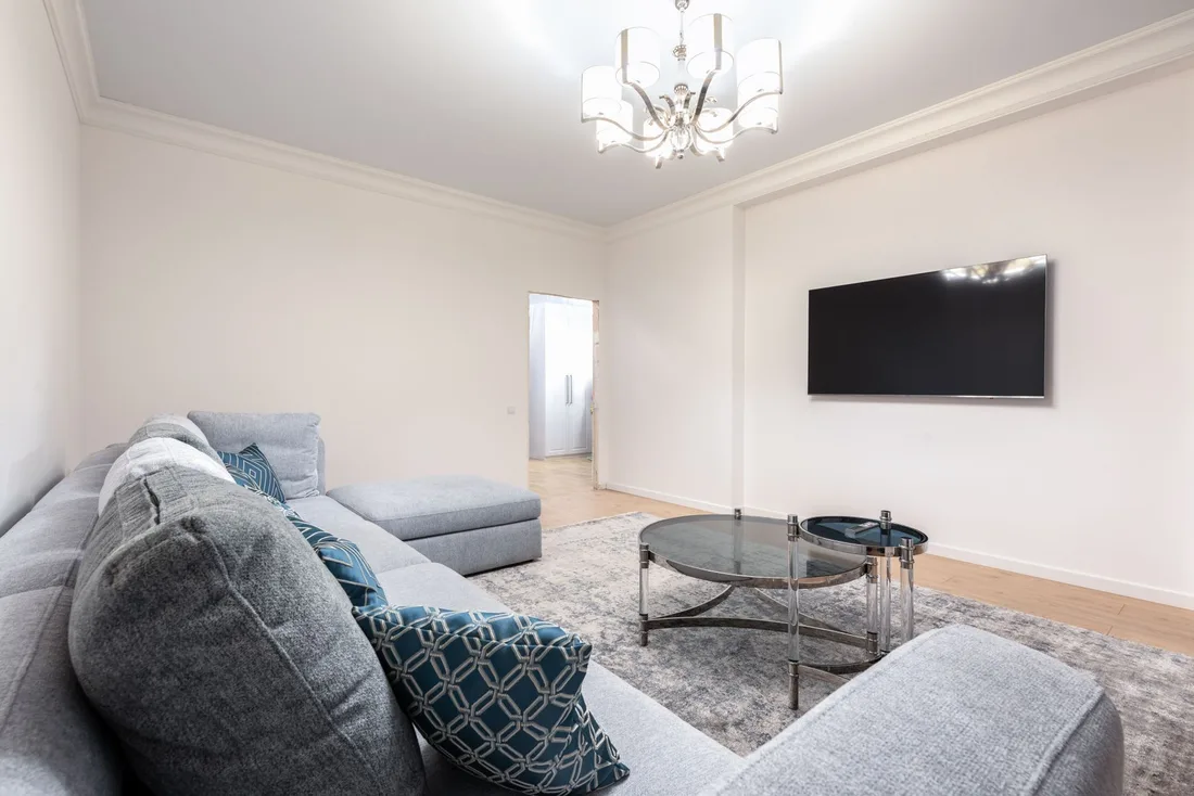 Restored bright living room with grey sofa, wall-mounted TV and chandelier. Photo by Max Vakhtbovych on Pexels