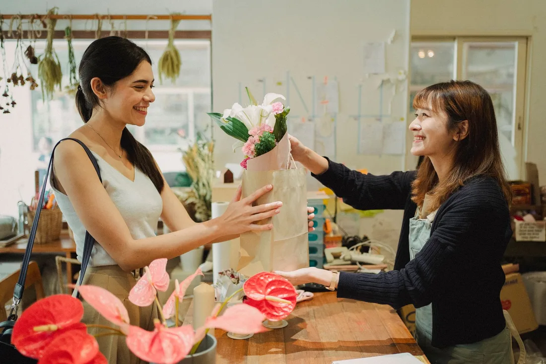 Side view of cheerful florist in apron giving paper bag with bouquets of flowers to smiling customer in floral shop in daytime. Photo by Amina Filkins on Pexels