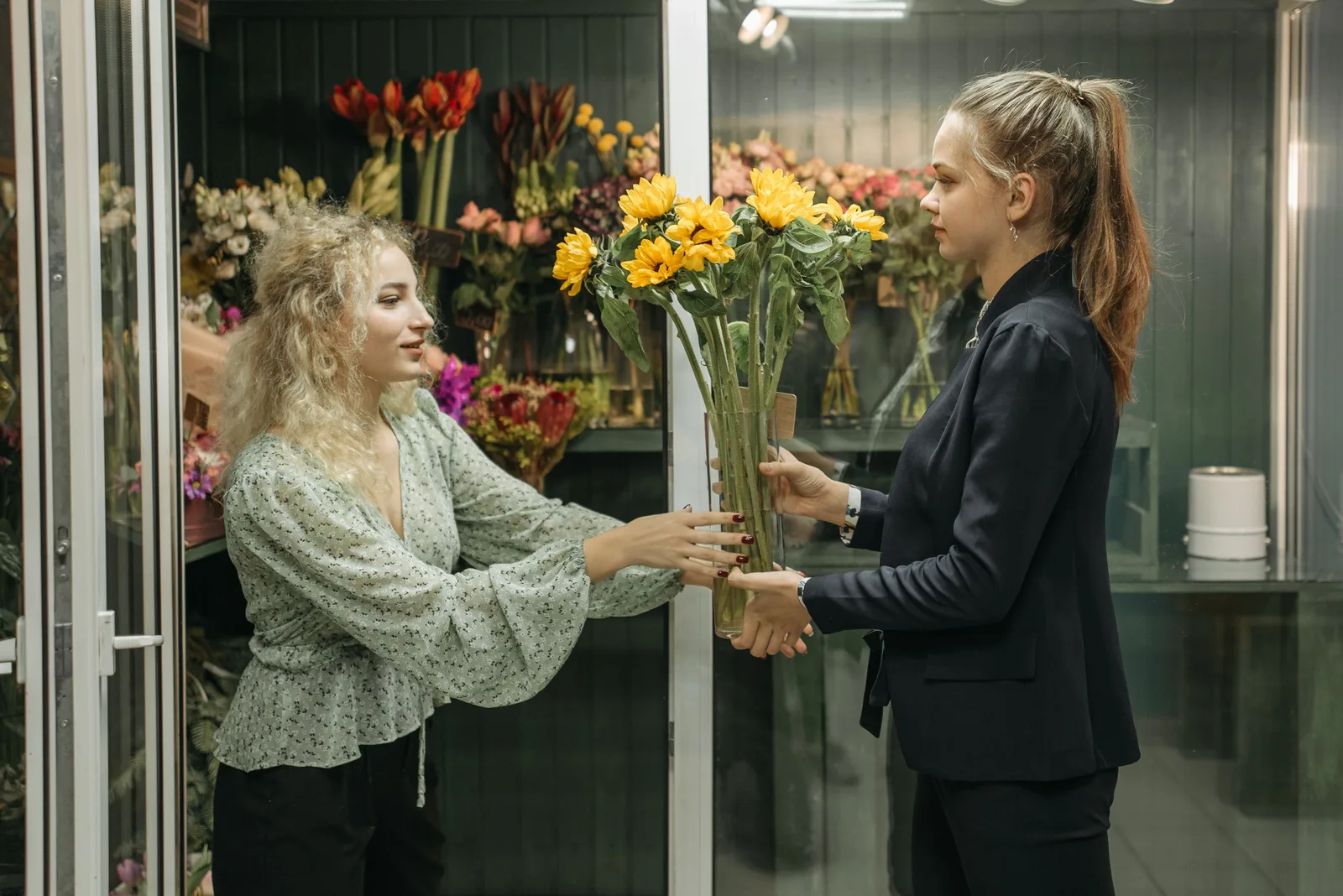 Two women standing, handing over sunflowers in a vibrant flower shop setting. Photo by Pavel Danilyuk on Pexels
