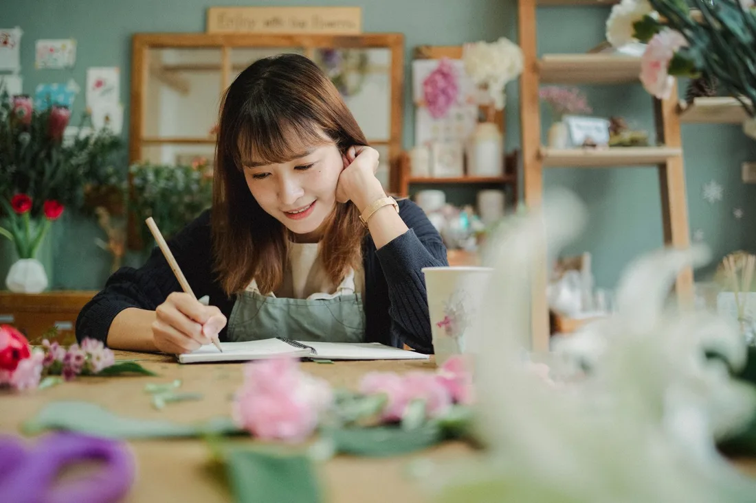Smiling young female florist in apron writing notes while working in cozy flower shop. Photo by Amina Filkins on Pexels