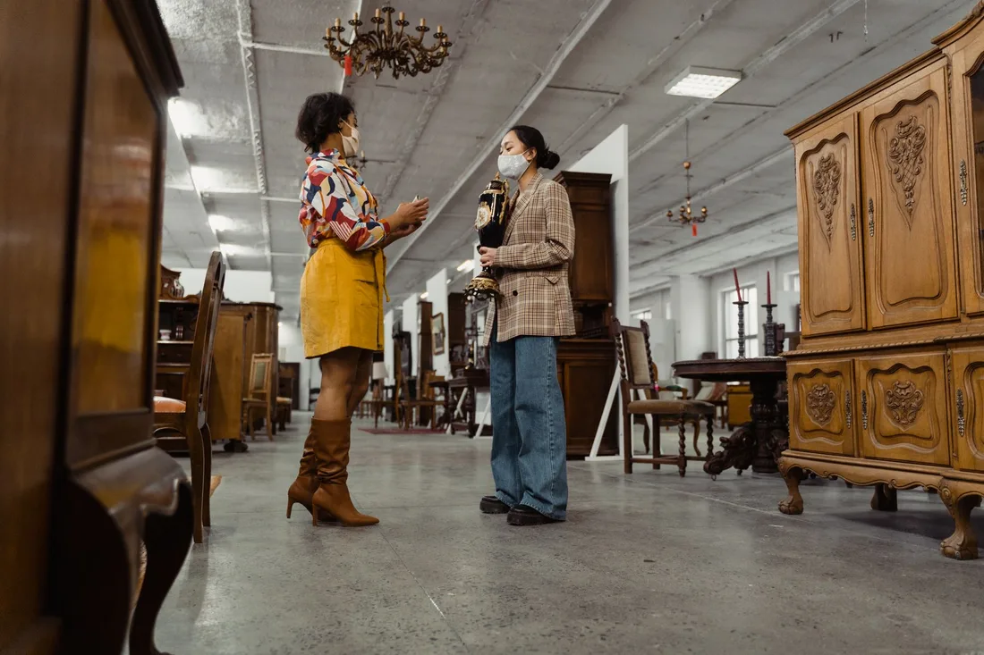 Two women wearing masks browsing antique furniture in a vintage store setting. Photo by Tima Miroshnichenko on Pexels