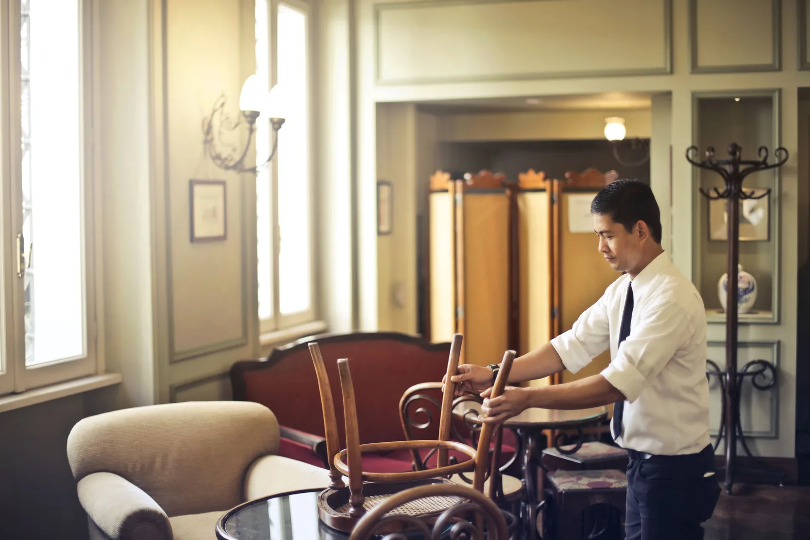Side view of Asian male employee in formal wear standing near table and neatly stacking wooden chair on table in closed restaurant. Photo by Andrea Piacquadio on Pexels
