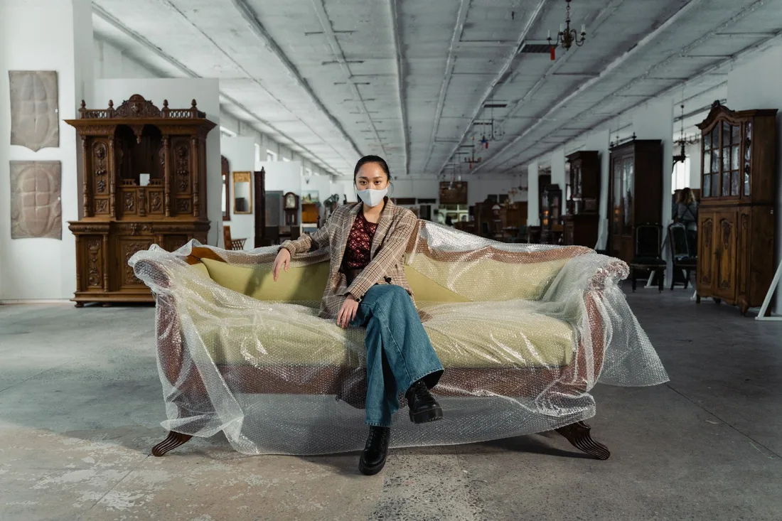 A woman in a mask sits on a wrapped sofa in an antique shop, surrounded by vintage furniture. Photo by Tima Miroshnichenko on Pexels