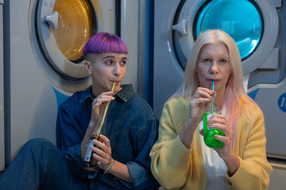 Two women relaxing with drinks beside colorful washing machines in a laundromat. Photo by cottonbro studio on Pexels