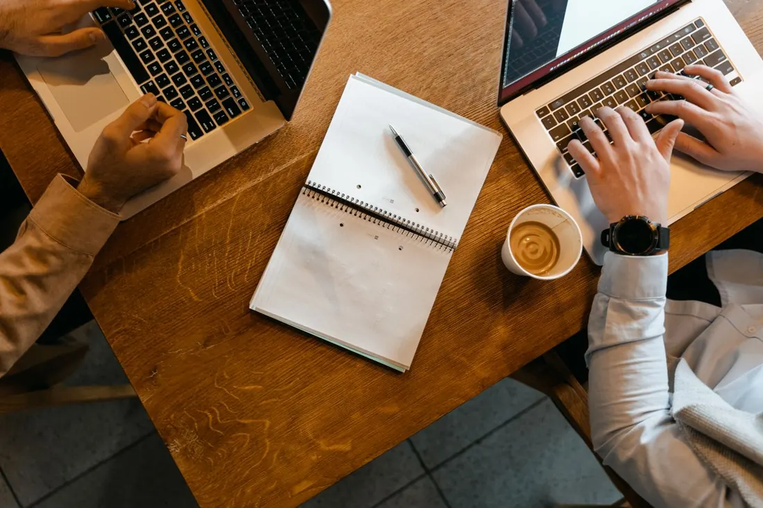 Two people typing on laptops at a wooden table, with a notebook, a pen and a coffee cup between them. Photo by MART PRODUCTION on Pexels