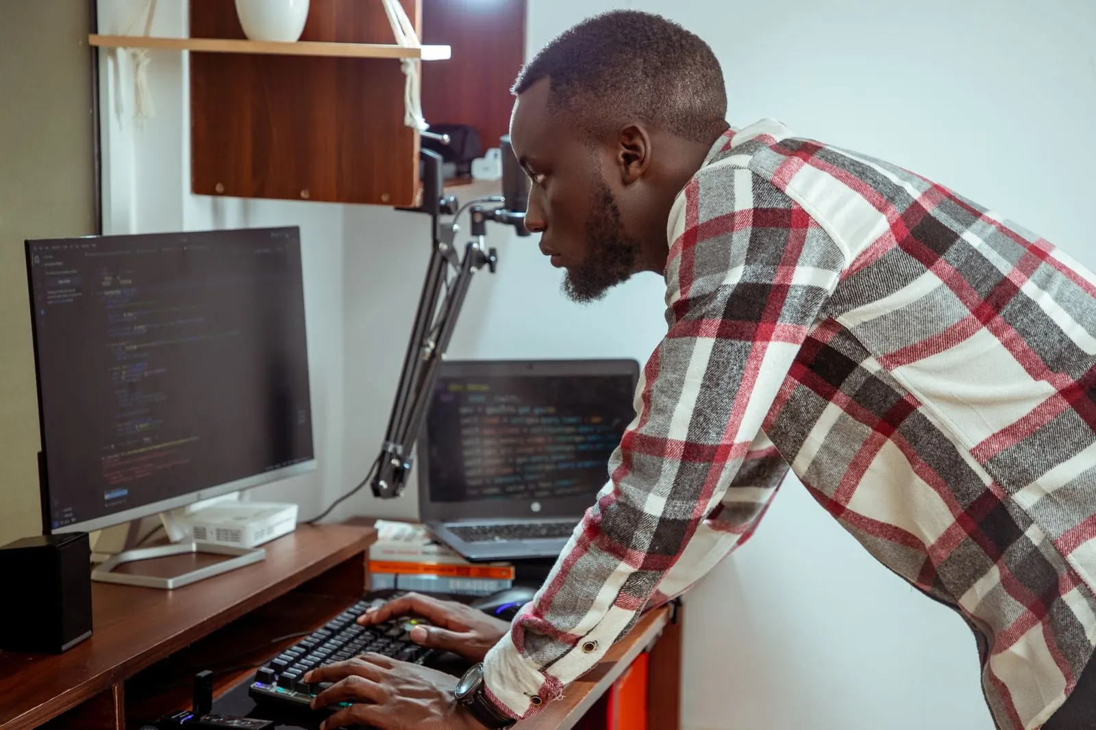 IT specialist leaning over a desk, typing while a desktop monitor and a laptop both show code. Photo by Naboth Otieno on Pexels