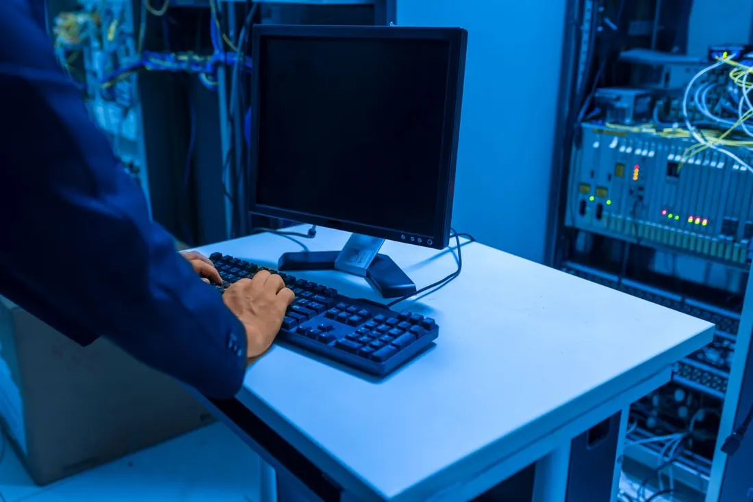 A technician at a keyboard in a server room, with equipment racks and indicator lights beside the desk. Photo by panumas nikhomkhai on Pexels