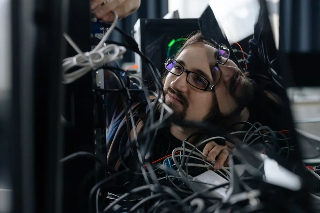 A technician sorting a tangle of cables behind a desktop computer. Photo by cottonbro studio on Pexels