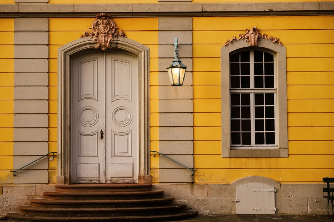 Ornate white front door of a yellow house with stone steps and a lantern. Photo by Christina & Peter on Pexels