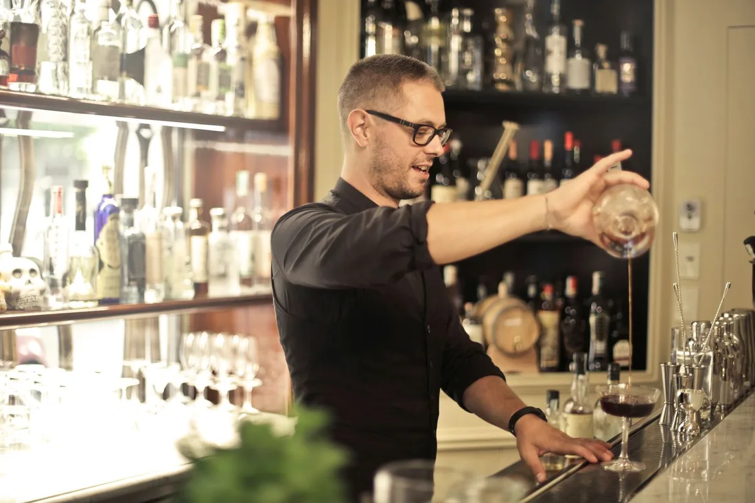 Side view of positive young barman in black uniform and eyeglasses pouring wine into glass while working at bar counter in modern restaurant. Photo by Andrea Piacquadio on Pexels